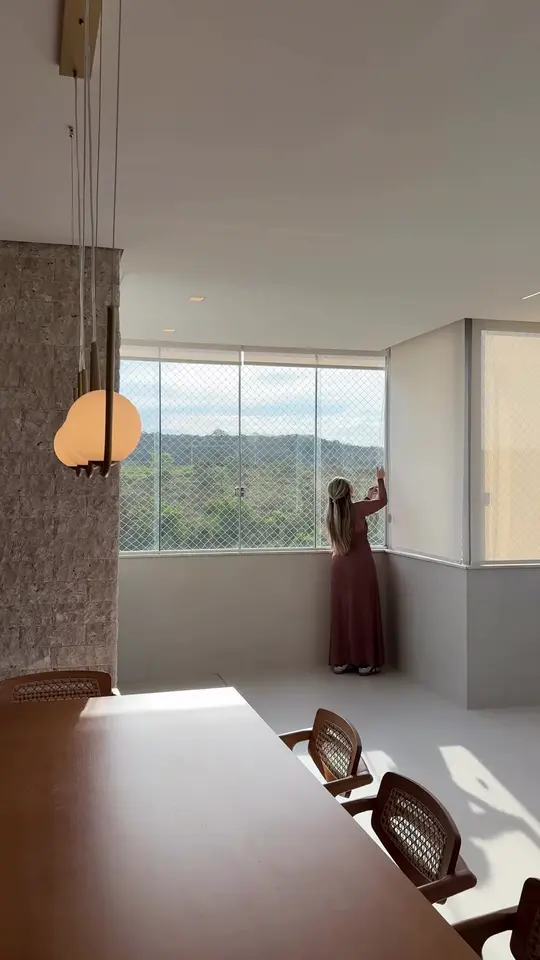 Sala ampla com janelas panorâmicas voltadas para a mata, onde as persianas rolô são baixadas uma a uma